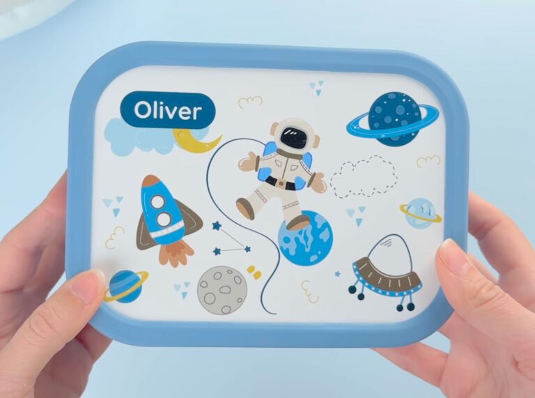 Matboks og drikkeflaske til barn -En blå matboks med navn og astronautdesign, holdt av en person.