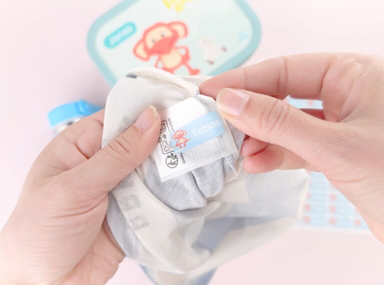 Navnelapp som plasseres på vaskeanvisningen inni et barneplagg. I bakgrunnen ser man en personlig matboks med Fantorangen.