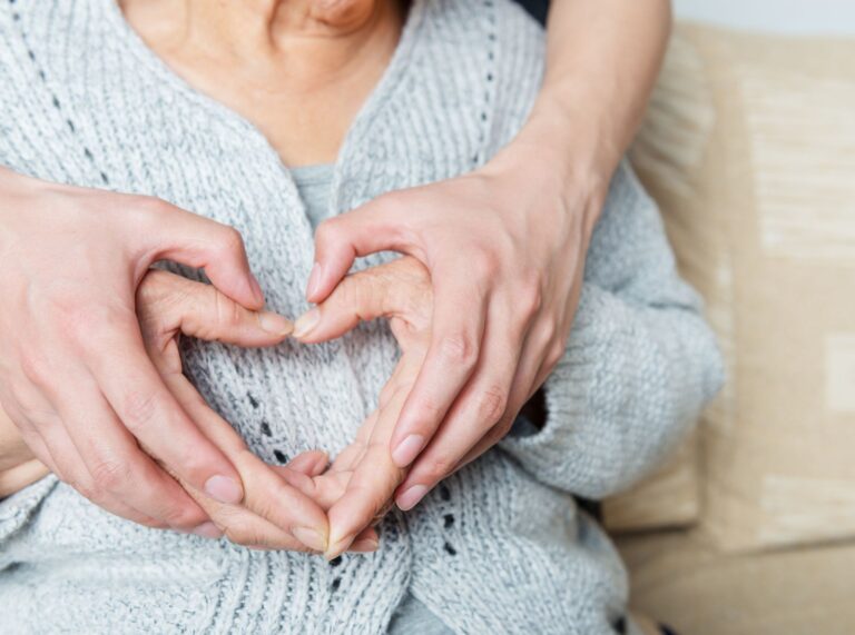 En eldre person i ullgenser sitter med hendene formet som et hjerte. En yngre person holder hendene over og fullfører hjerteformen.