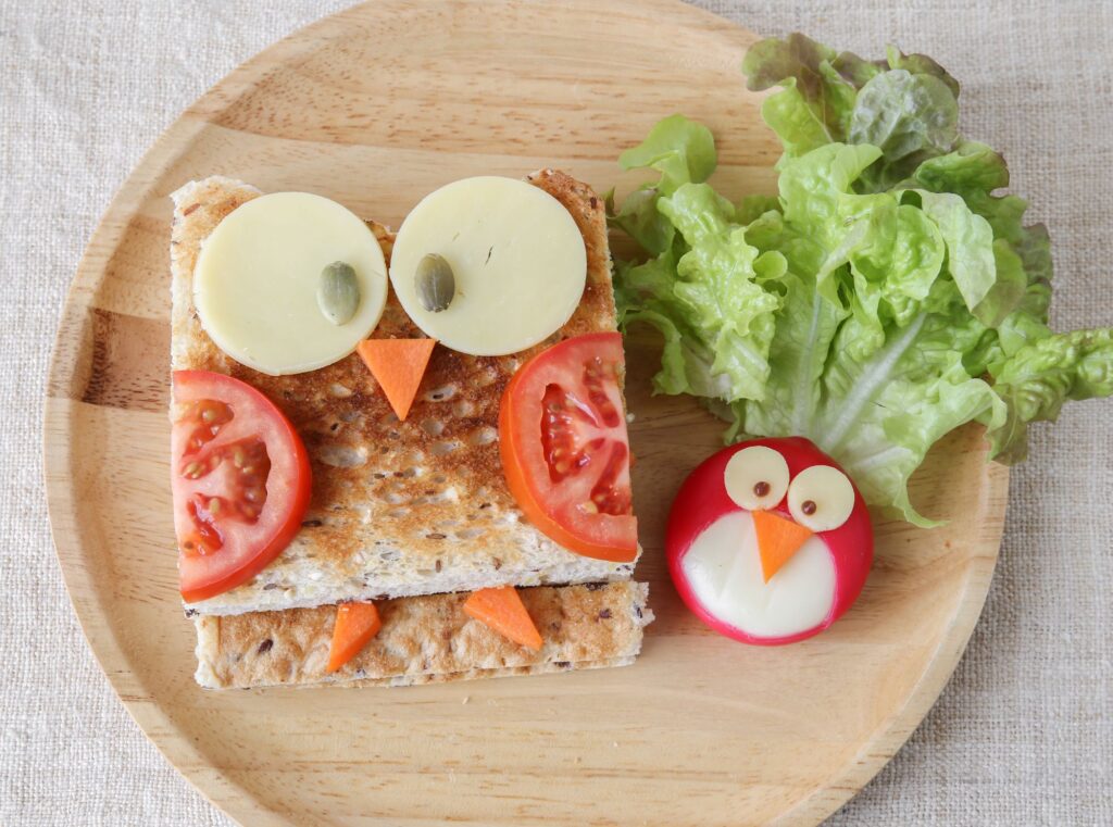 En matpakke med brødskive, frukt og grønnsaker, servert på en tre-tallerken. Maten er kuttet og tilberedt på en kreativ måte, slik at den danner to søte uglefigurer
